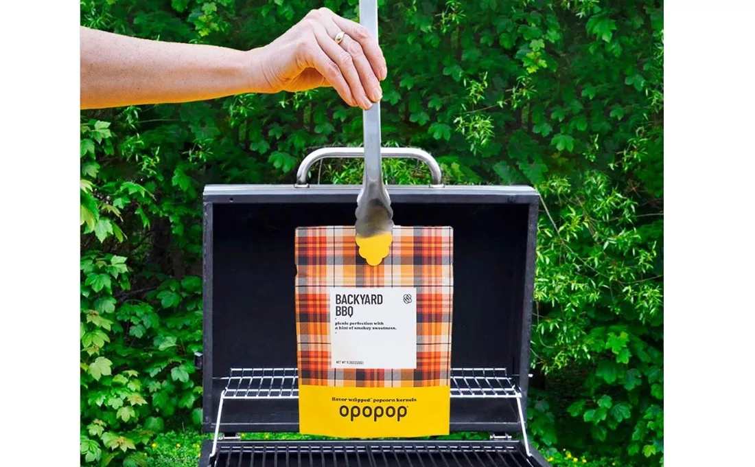 Hand holding tongs with Opopop popcorn bag over grill