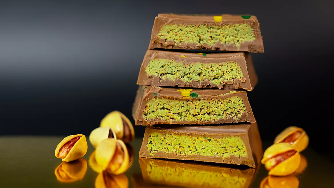 A tower of four Dubai chocolate halves with kataifi dough and emerald pistachio paste, whole pistachios with a dark gray background.