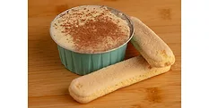 Ramekin of tiramisu next to ladyfingers on wood table