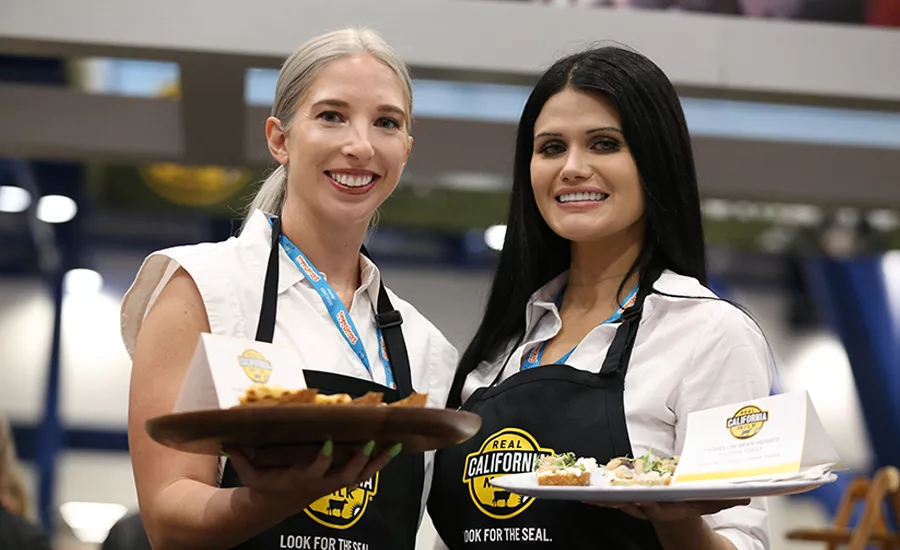 Two women offer samples of bread and cheese on a tray.