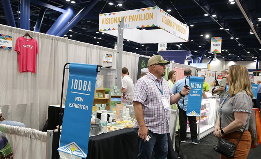 A man and a woman talk in front of a sign that says IDDBA New Exhibitor.