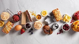 This image displays a collection of ingredients and finished baked goods, including cupcakes, pastries, fresh fruit, jams, and baking tools. 