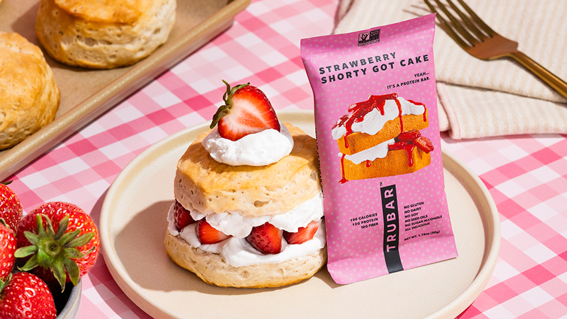 Trubar’s Strawberry Shorty Got Cake bar in pink polka dot packaging on a plate next to a strawberry shortcake biscuit plus a bowl of strawberries and some biscuits.