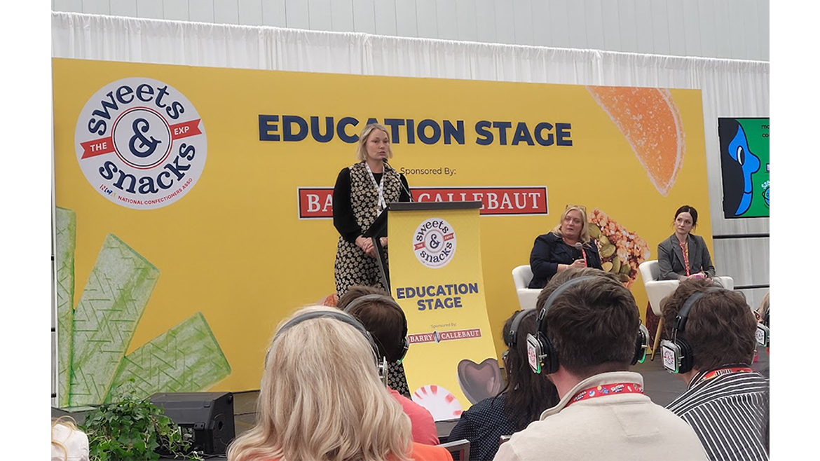 A woman presents information at the 2025 Sweets & Snacks Expo Education stage, with two other women listening to her on stage and audience members wearing headphones.