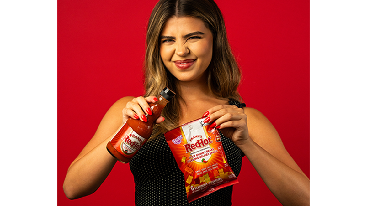 A woman with brown and blonde hair with a V-neck polka dot shirt holds a bottle of Frank’s RedHot in one hand and a bag of Frank’s RedHot and Huer Foods Gummies in the other.