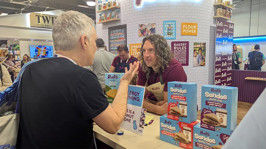 Man with long wavy hair at Rudi’s booth talking to gray-haired man
