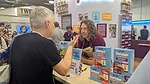 Man with long wavy hair at Rudi’s booth talking to gray-haired man