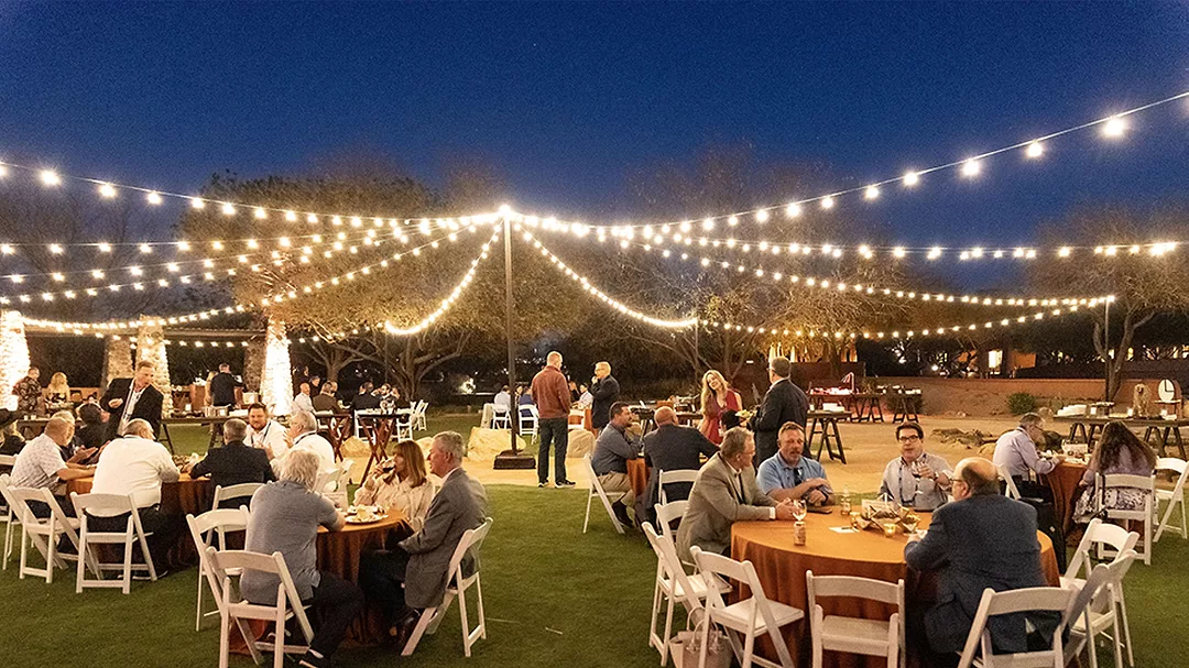 This image captures an outdoor evening reception, likely a corporate holiday party or a wedding, featuring festive string lights overhead.