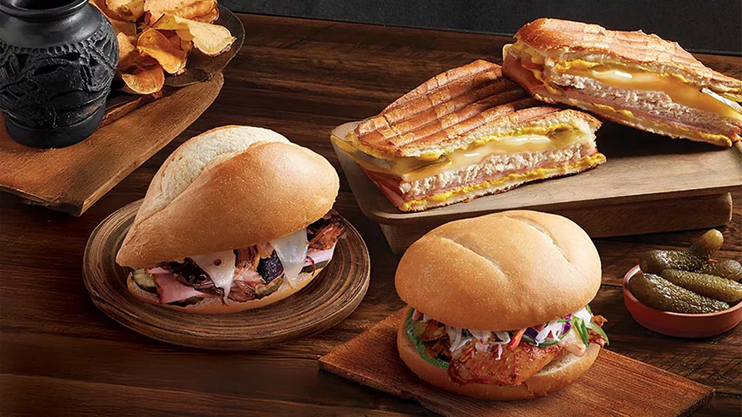 This image shows a variety of artisanal sandwiches, likely featuring a traditional pressed Cuban sandwich (Cubano) which is made with ham, roasted pork, Swiss cheese, pickles, and mustard.