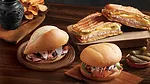 This image shows a variety of artisanal sandwiches, likely featuring a traditional pressed Cuban sandwich (Cubano) which is made with ham, roasted pork, Swiss cheese, pickles, and mustard. 