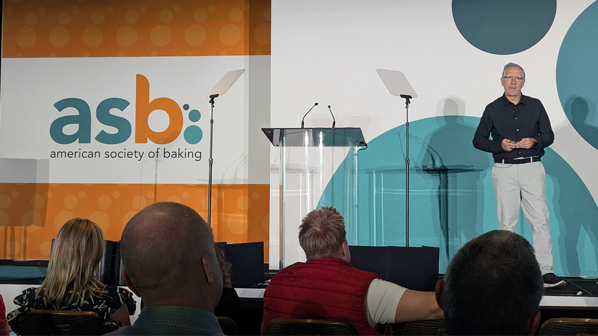 A man stands on a stage with the ASB American Society of Baking logo and branding behind him to the left.