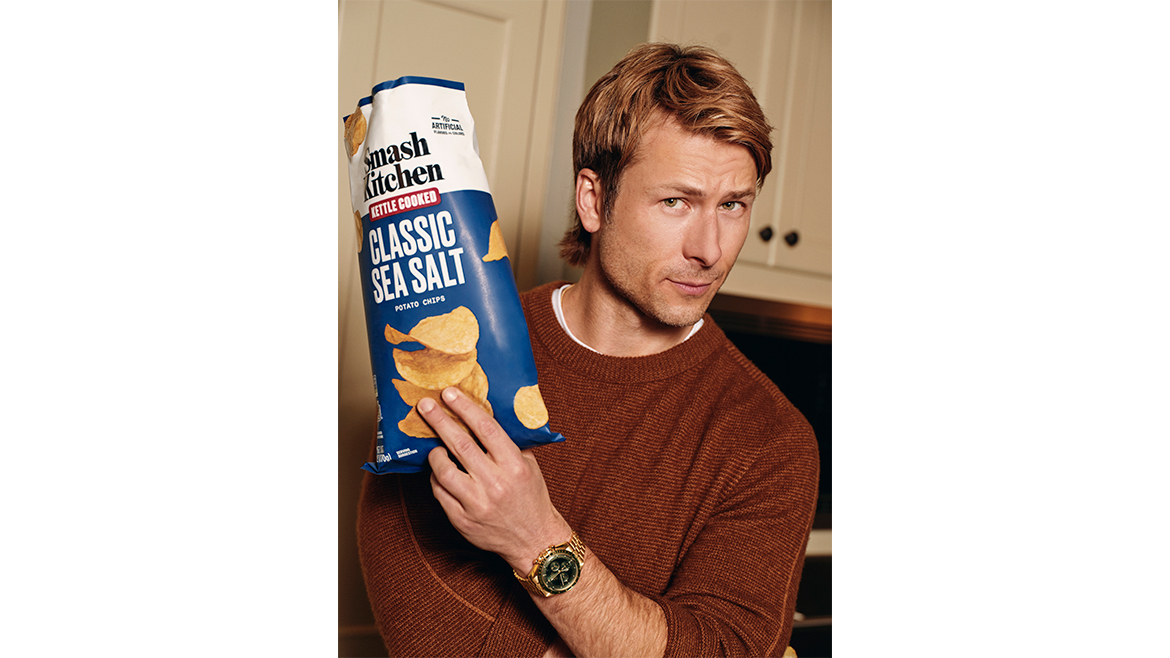 Actor Glen Powell holding up a blue and white bag of Smash Kitchen Classic Sea Salt chips.