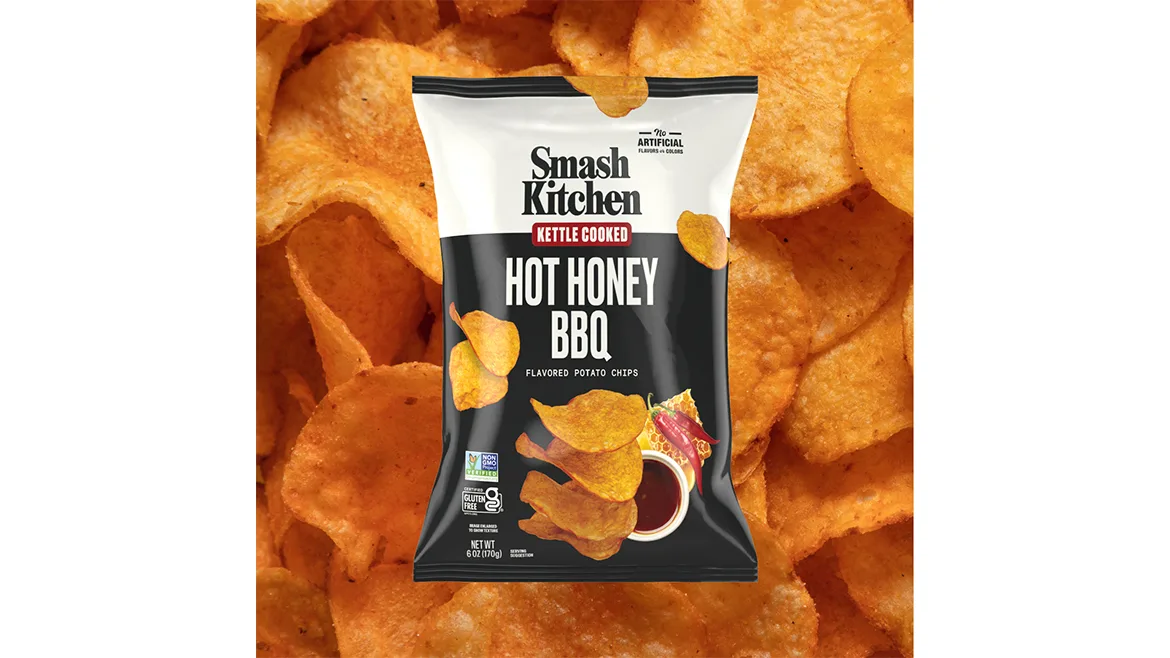 A black and white bag of Smash Kitchen Kettle Cooked Hot Honey BBQ chips, with chips in the background. A black and white bag of Smash Kitchen Kettle Cooked Hot Honey BBQ chips, with chips in the background.