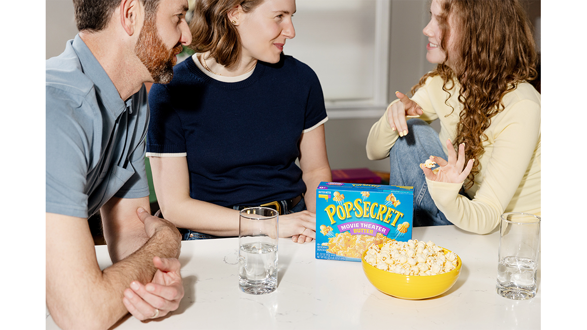 a family sitting at a table with a box of Pop Secret Microwave Popcorn. 