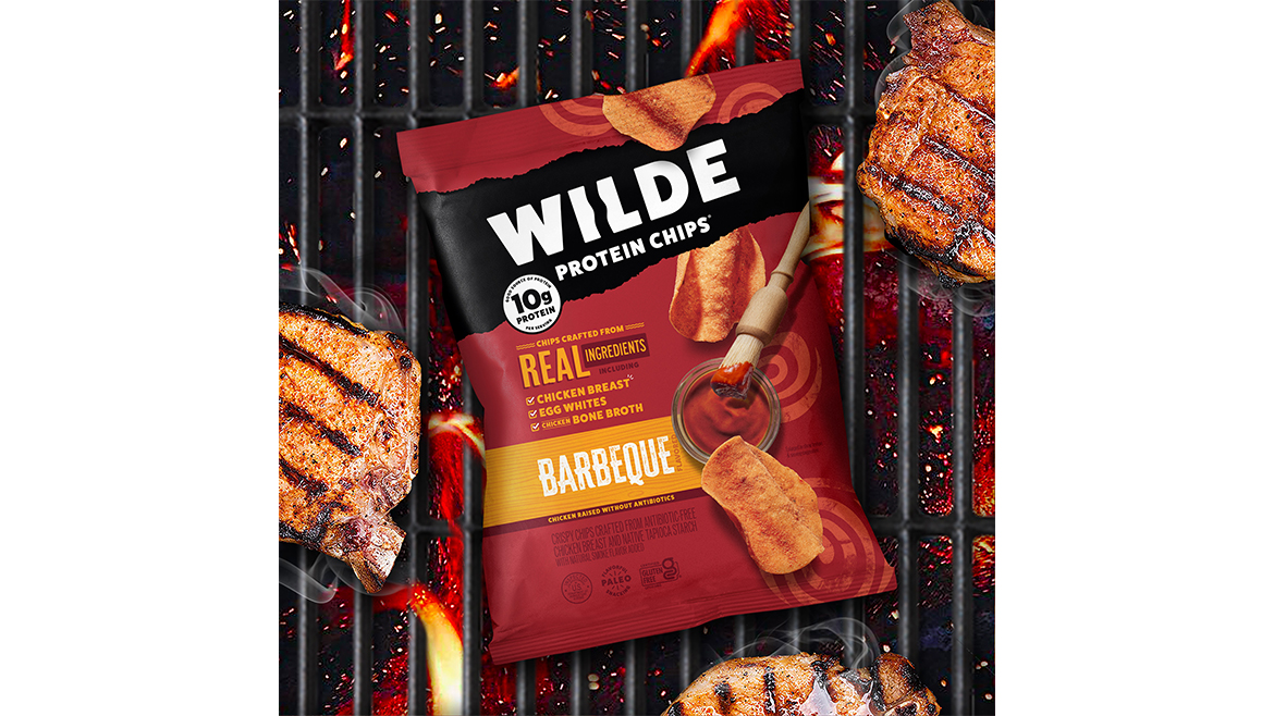 A red bag of Wilde Protein Chips Barbecue flavor, on top of a grill with steaks cooking around it.