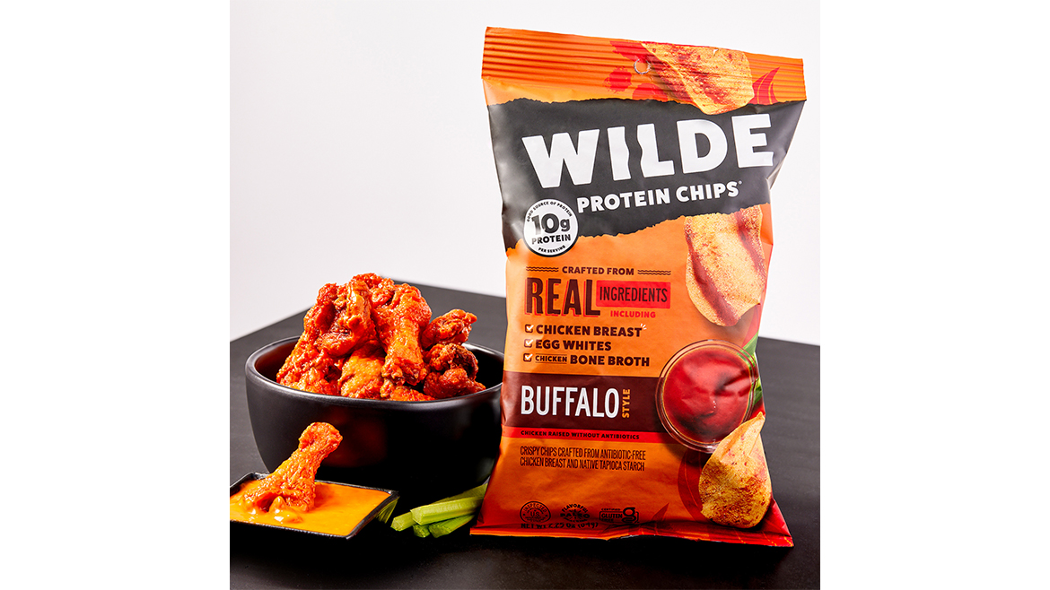 An orange bag of Wilde Protein Chips Buffalo flavor, next to a bowl with buffalo wings stacked in it, plus one in dipping sauce, and celery.