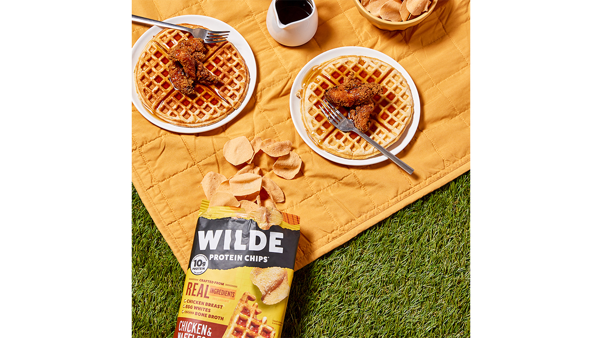 An open, yellow bag of Wilde Chicken and Waffles protein chips, spilling out onto a picnic blanket with two plates of waffles topped with chicken next to it.