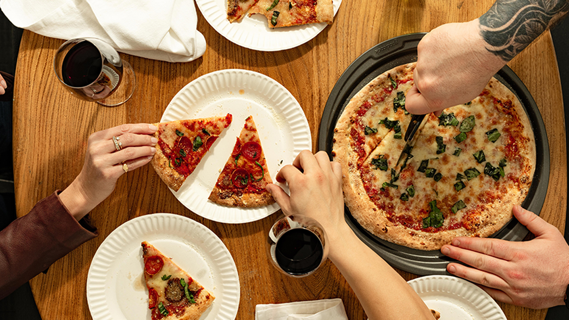 Hands and arms reaching for pizza slices with one person with an arm tattoo cutting a pizza with a pizza cutter.