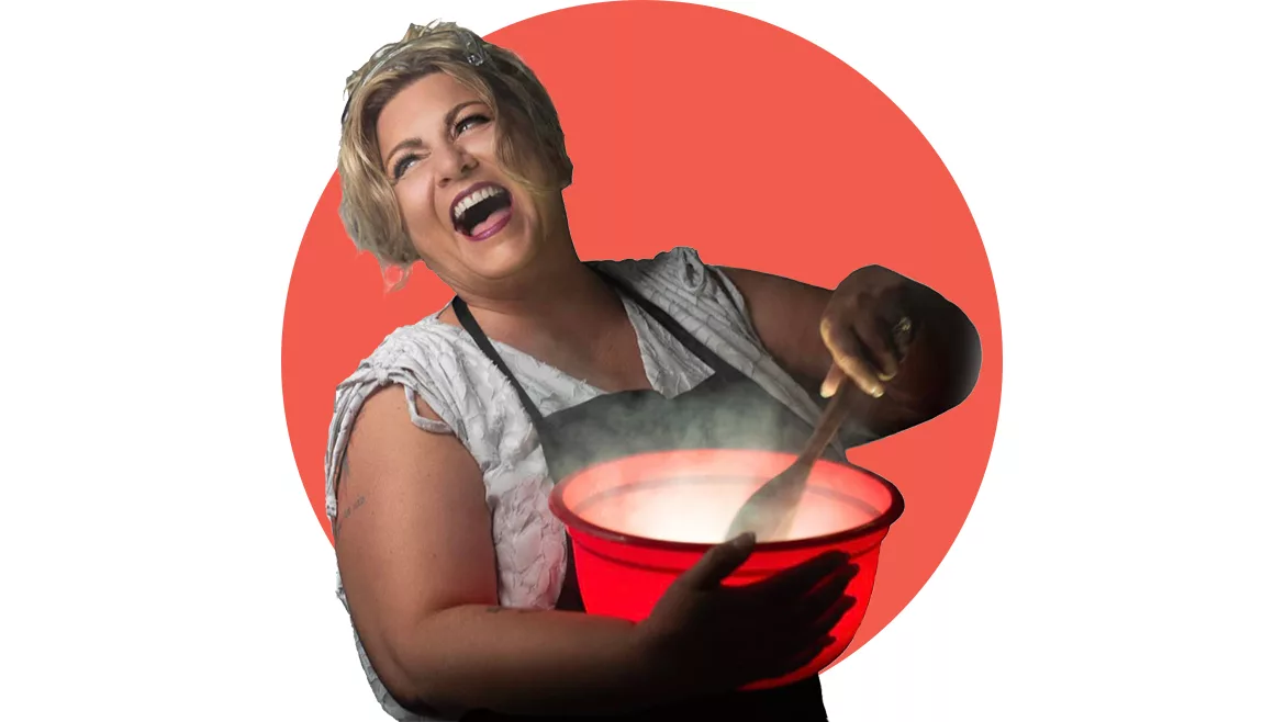 White woman with short blonde hair in blouse and apron holding steaming red bowl in arm, stirring a spoon with the other, and laughing over her shoulder.