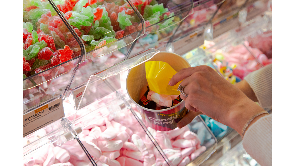 A mix of colorful Swedish candies in plastic boxes with a woman’s hand reaching in with a yellow scooper.