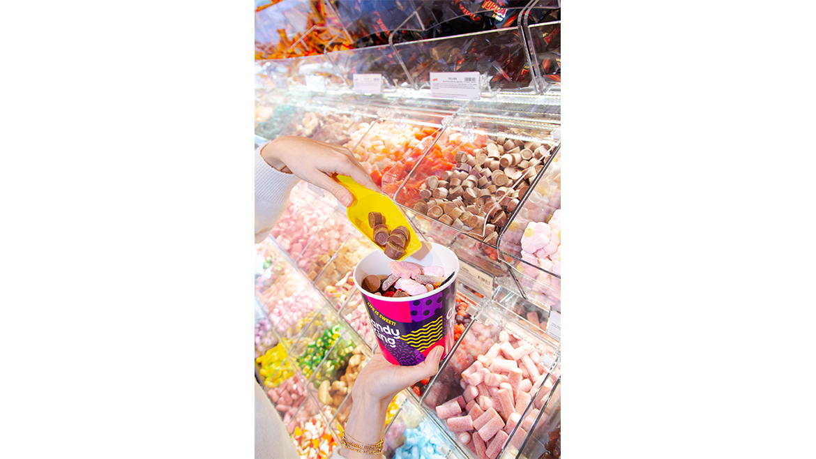A mix of colorful Swedish candies in plastic boxes with a woman’s hand reaching in with a yellow scooper.