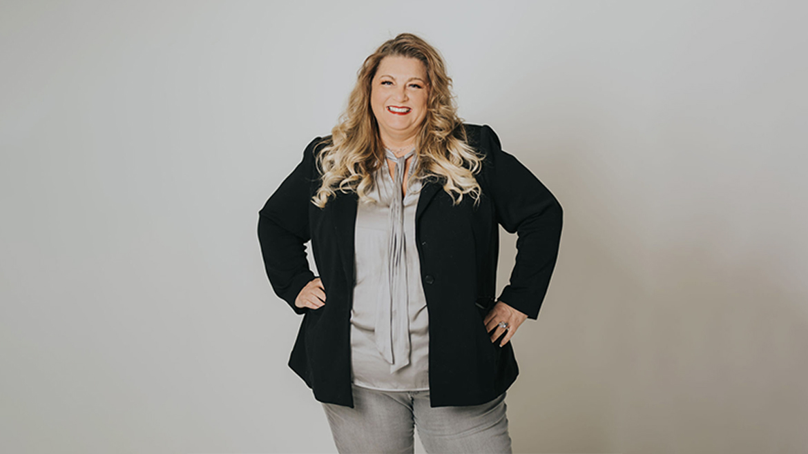 Professional photo of white woman with long blonde hair, in black blazer and light colored blouse and khakis, standing with hands on hips and smiling.