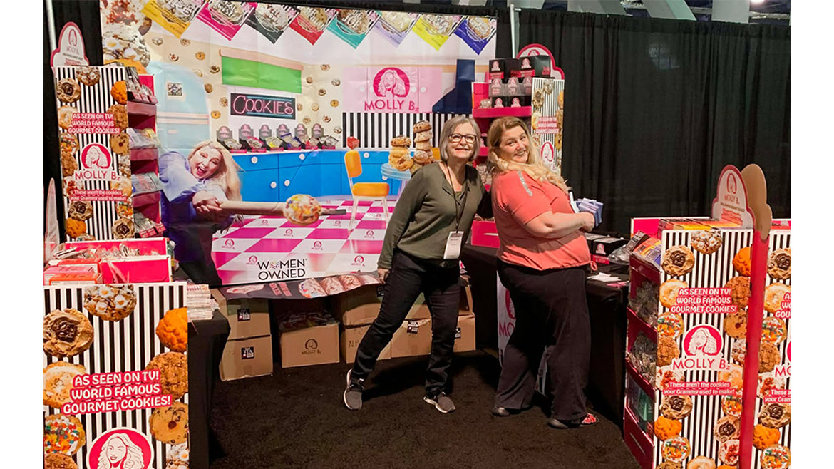 Two women in colorful tradeshow booth surrounded by cookies