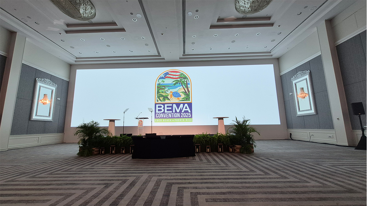 BEMA Convention 2025 on a large screen, with three podiums in front of it and a long table.