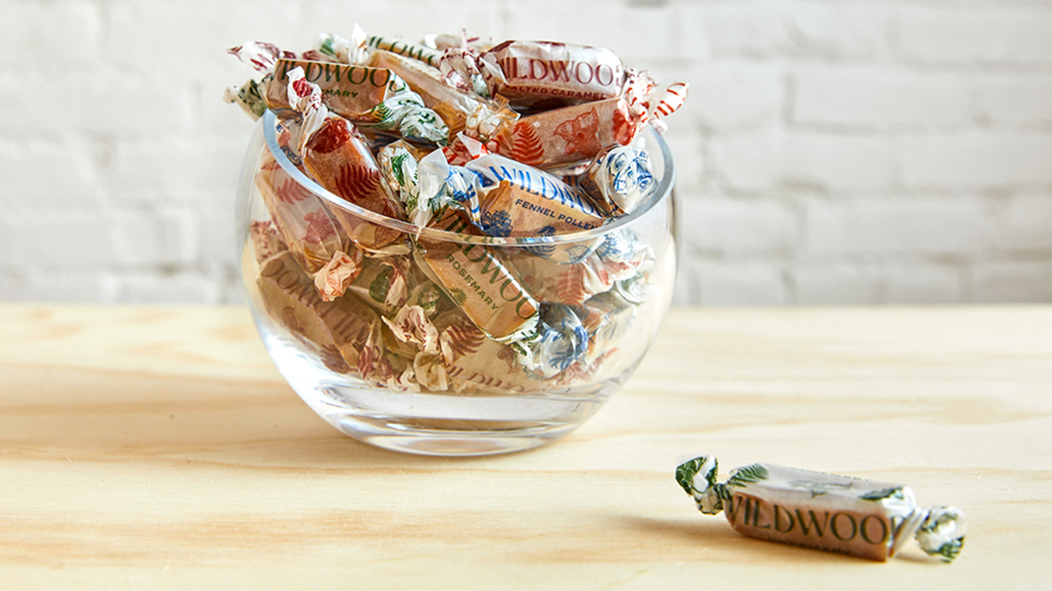 A glass bowl on a flat wood surface filled with individually wrapped Wildwood caramels.