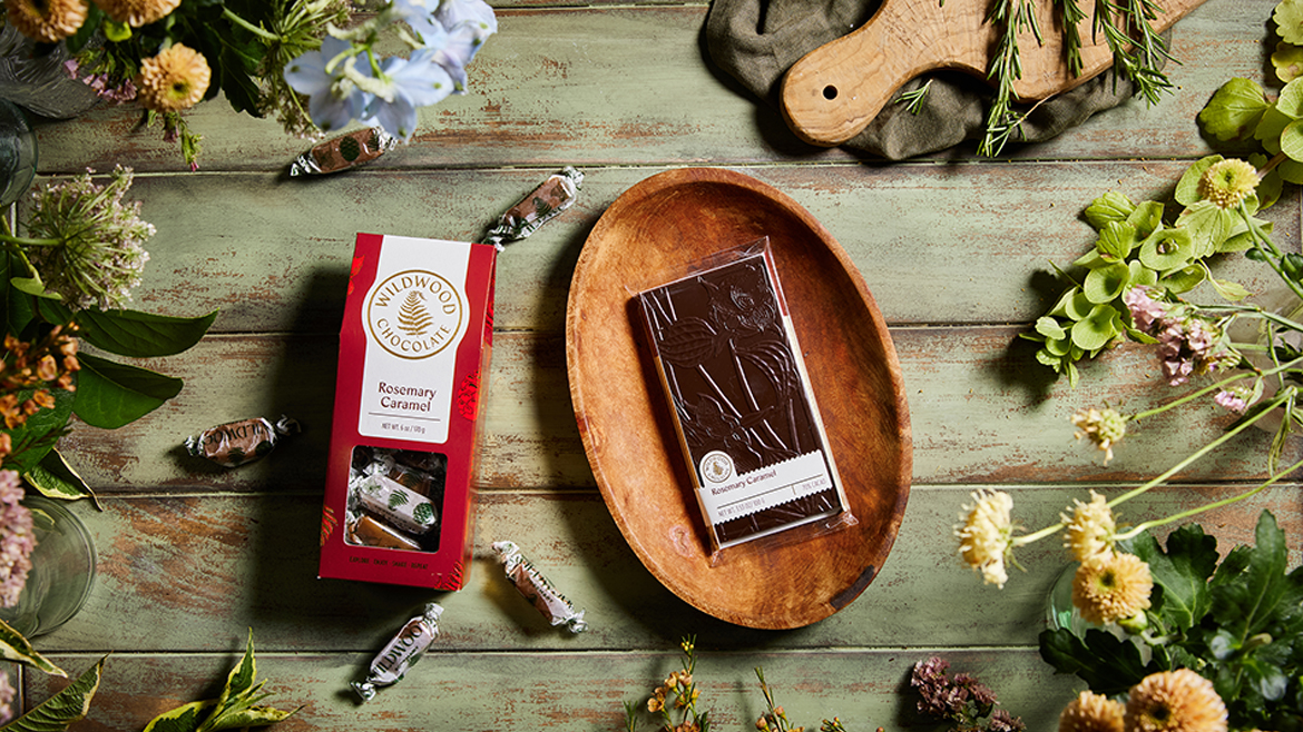 A red package of Wildwood’s Rosemary Caramel next to a shallow wooden bowl with its Rosemary Caramel chocolate, against a background of distressed wood, plus flowers.